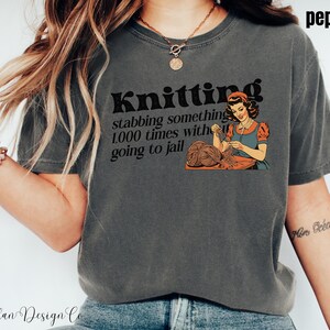 May include: A gray t-shirt with a vintage-style graphic that says "Knitting stabbing something 1,000 times with going to jail". The graphic features a woman knitting.