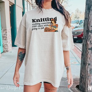 May include: A woman wearing a light beige t-shirt with the text "Knitting stabbing something 1000 times without going to jail" and a cartoon image of a woman knitting.