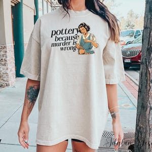 May include: A light beige t-shirt with a graphic of a woman making pottery. The text on the shirt reads "pottery because murder is wrong".