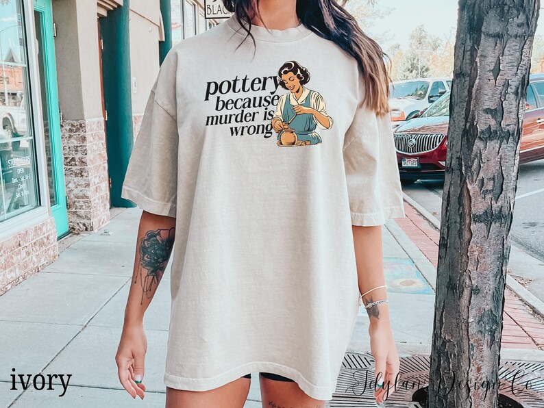 May include: A light beige t-shirt with a graphic of a woman making pottery. The text on the shirt reads "pottery because murder is wrong".