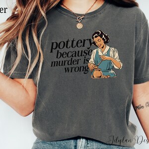 May include: A dark gray t-shirt with the text "pottery because murder is wrong" and a cartoon image of a woman making pottery.