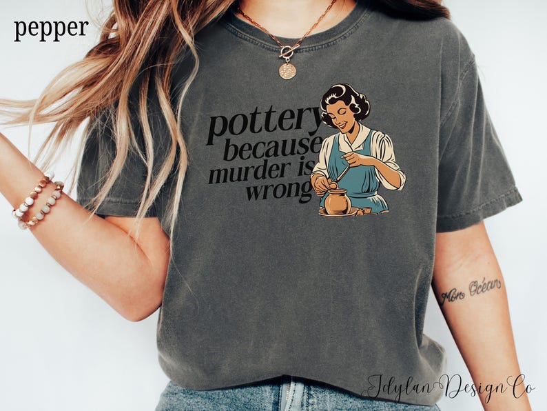 May include: A dark gray t-shirt with the text "pottery because murder is wrong" and a cartoon image of a woman making pottery.