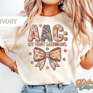 May include: Ivory-colored t-shirt with the text "AAC: MY FIRST LANGUAGE." The letters are decorated with floral patterns. A large bow with floral designs is below the text, surrounded by small flowers. The shirt has short sleeves.