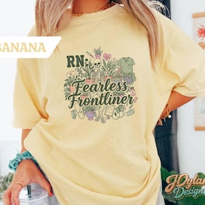 May include: A light yellow t-shirt with the text "RN: Fearless Frontliner" and a graphic of medical and plant-related elements. The shirt has short sleeves and a relaxed fit.