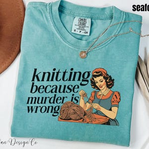Op de afbeelding: Zeegroen t-shirt met de tekst "knitting because murder is wrong" en een vintage illustratie van een breiende vrouw. Het shirt heeft een ronde hals en korte mouwen. Een gouden ketting met een hanger ligt op het shirt.