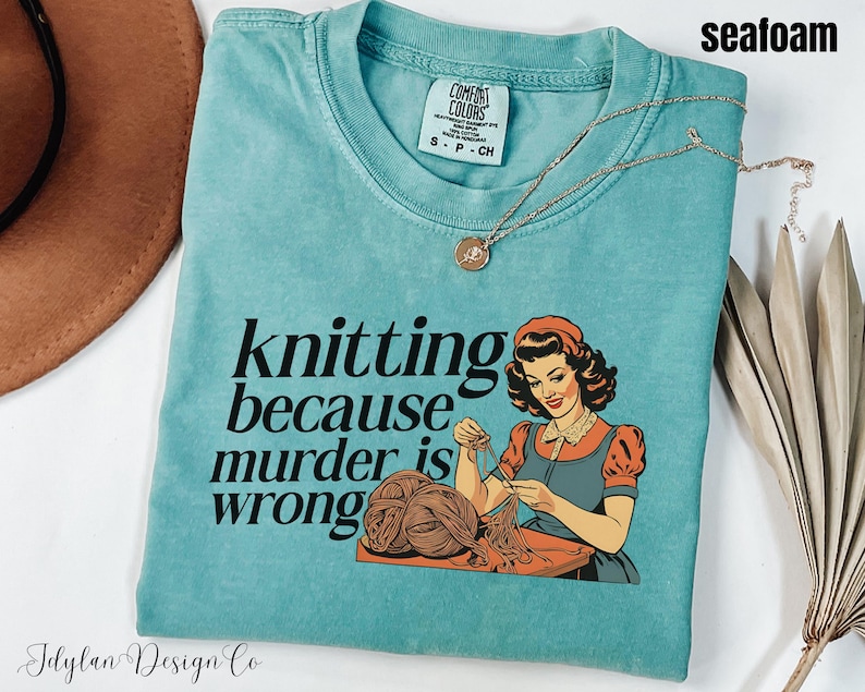 Op de afbeelding: Zeegroen t-shirt met de tekst "knitting because murder is wrong" en een vintage illustratie van een breiende vrouw. Het shirt heeft een ronde hals en korte mouwen. Een gouden ketting met een hanger ligt op het shirt.