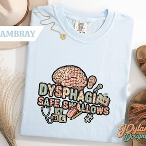 May include: Light blue t-shirt with a graphic design featuring a brain, ear, microphone, and other medical symbols. The text "DYSPHAGIA SAFE SWALLOWS" is prominently displayed. The shirt is a Comfort Colors brand.