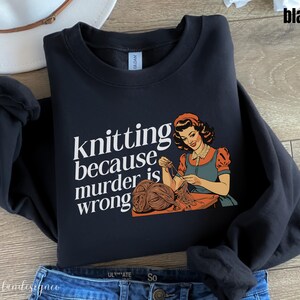 Op de afbeelding: Zwart sweatshirt met de tekst "knitting because murder is wrong" en een vintage illustratie van een breiende vrouw. De vrouw draagt een oranje jurk en een blauw schort. Het sweatshirt wordt gecombineerd met een blauwe jeans.