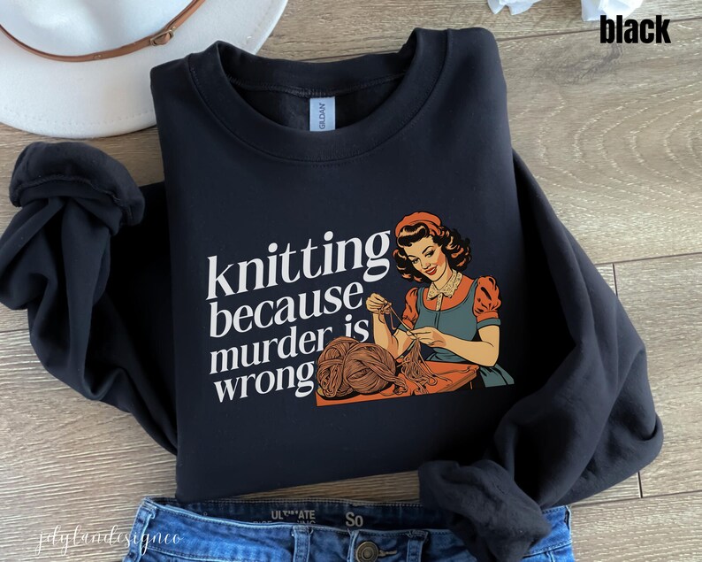 Op de afbeelding: Zwart sweatshirt met de tekst "knitting because murder is wrong" en een vintage illustratie van een breiende vrouw. De vrouw draagt een oranje jurk en een blauw schort. Het sweatshirt wordt gecombineerd met een blauwe jeans.