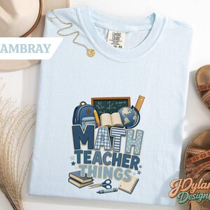 May include: Light blue t-shirt with a graphic that says "MATH TEACHER THINGS." The graphic includes a backpack, books, a globe, and school supplies in shades of blue. The shirt is a Comfort Colors brand.