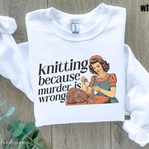 Op de afbeelding: Witte sweatshirt met de tekst "knitting because murder is wrong" en een vintage illustratie van een breiende vrouw. De vrouw draagt een rode en blauwe jurk en werkt met garen. De sweatshirt heeft lange mouwen.