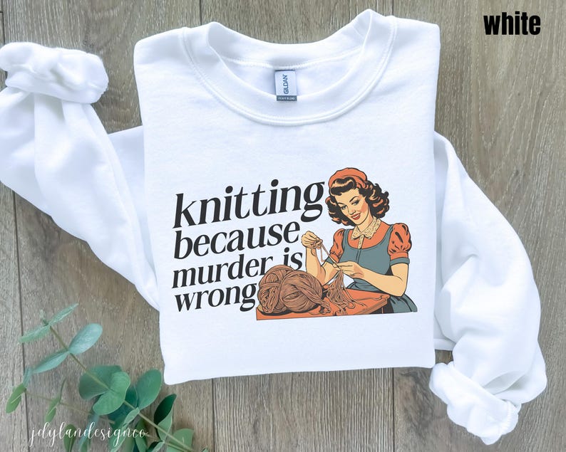 Op de afbeelding: Witte sweatshirt met de tekst "knitting because murder is wrong" en een vintage illustratie van een breiende vrouw. De vrouw draagt een rode en blauwe jurk en werkt met garen. De sweatshirt heeft lange mouwen.