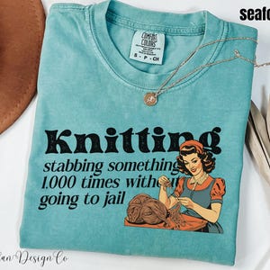May include: Seafoam blue t-shirt with a vintage-style graphic that says "Knitting stabbing something 1,000 times without going to jail". The graphic features a woman knitting.