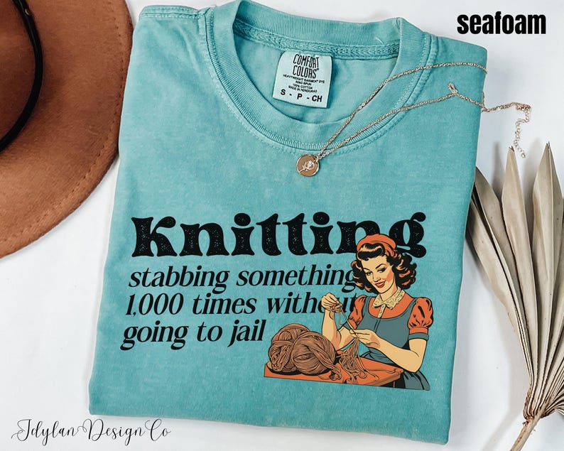 May include: Seafoam blue t-shirt with a vintage-style graphic that says "Knitting stabbing something 1,000 times without going to jail". The graphic features a woman knitting.