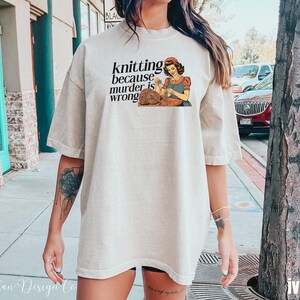 Op de afbeelding: Ivoor kleurig t-shirt met de tekst "knitting because murder is wrong" en een illustratie van een breiende vrouw. Het shirt heeft een relaxte pasvorm en korte mouwen. Comfortabele, casual kleding.