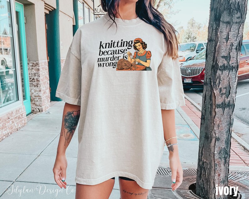 Op de afbeelding: Ivoor kleurig t-shirt met de tekst "knitting because murder is wrong" en een illustratie van een breiende vrouw. Het shirt heeft een relaxte pasvorm en korte mouwen. Comfortabele, casual kleding.