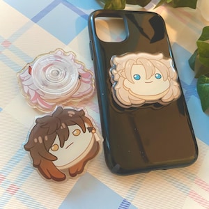 Könnte beinhalten: Drei transparente Handy-Anhänger aus Acryl mit Comicfiguren. Die Anhänger sind an einer schwarzen Handyhülle befestigt. Die Figuren sind ein weißhaariger Charakter mit blauen Augen, ein braunhaariger Charakter mit gelben Augen und ein weißer Charakter mit einem durchsichtigen Plastikring.