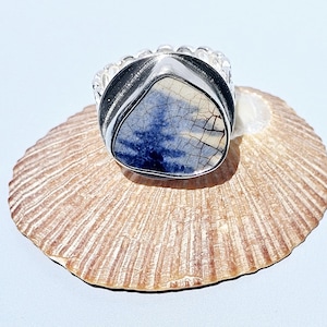 May include: A silver ring with a large, triangular stone featuring a blue and cream-colored pattern. The ring is set on a textured, tan seashell, creating a contrast of textures and colors. The ring's band has a decorative, ridged design.