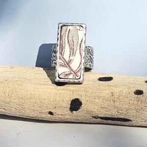 Pode incluir: Anel retangular com um design floral branco e marrom, emoldurado em uma estrutura de metal prateado. A banda do anel tem um padrão texturizado. O anel é exibido em um pedaço de madeira flutuante contra um fundo azul claro.