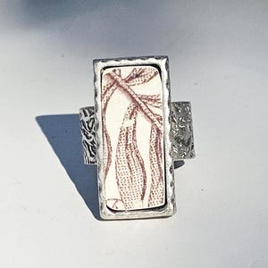 May include: A silver-toned ring with a rectangular, off-white ceramic centerpiece featuring a brown botanical pattern. The ring has a textured band and a hammered bezel around the ceramic. The design is vintage-inspired.