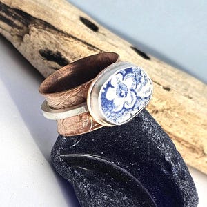 May include: A stack of handcrafted rings. The top ring features a blue and white floral design set in a silver bezel. Below, a copper band with an embossed pattern sits atop a silver band and a copper ring.