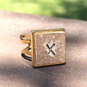 May include: A gold ring with a square-shaped stone featuring a textured surface and a black 'X' etched into the center.