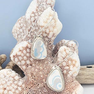 May include: A pair of silver dangle earrings. Each earring features a teardrop-shaped, white ceramic piece with a blue and gray wave pattern, set in a silver bezel. The earrings are displayed on a piece of coral against a light blue background.