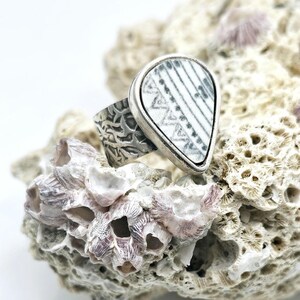 Könnte beinhalten: Ein Silberring mit einem tropfenförmigen, weiß-grau gemusterten Keramikeinsatz. Das Ringband hat ein strukturiertes Design. Der Ring wird auf einem Stück Koralle präsentiert.