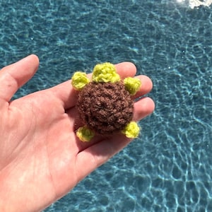 May include: A small crocheted turtle toy with a brown shell and green limbs. The turtle is being held in a hand against a blue background.