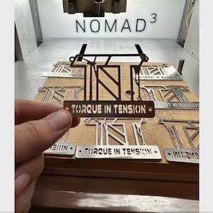 Könnte beinhalten: Ein Metallschild mit dem Text "TORQUE IN TENSION" wird vor einer Holzoberfläche mit anderen ähnlichen Schildern gehalten. Das Wort "NOMAD 3" ist im Hintergrund sichtbar. Die Schilder scheinen aus Metall geschnitten zu sein.