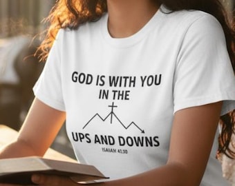 May include: White t-shirt with the text "GOD IS WITH YOU IN THE UPS AND DOWNS" and a mountain graphic. The shirt is worn by a person sitting at a wooden table, holding a book. A cup and a potted plant are also on the table.