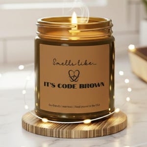 Puede incluir: Una vela encendida en un tarro de cristal con una etiqueta marrón que dice "Smells like... It's Code Brown". La vela está sobre un posavasos de madera. El fondo incluye una ventana, una planta pequeña y un libro.