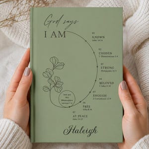 May include: A sage green hardcover book with the text God says I AM and a list of seven numbered spiritual affirmations including Known, Chosen, Strong, Beloved, Enough, Free, and At Peace, with the name Haleigh printed at the bottom.