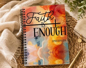 May include: Spiral-bound notebook with the words "Faith is Enough" and the Bible verse Ephesians 2:8. The cover features an abstract design in shades of orange, yellow, blue, and red. A gold pen and small beige wallet are also visible.
