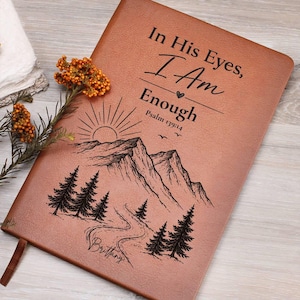 May include: A tan leather-like journal featuring the black engraved text In His Eyes, I Am Enough, Psalm 139:14, and the name Brittany. The cover displays a black line art illustration of a mountain range, a rising sun, pine trees, and a path.