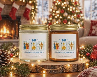 May include: Two glass jar candles with gold lids, one clear and one amber, featuring a white label with a blue bow, a nativity scene, and the text "OH COME LET US ADORE HIM" followed by "Eco-friendly | Non-toxic | Hand-poured in the USA".