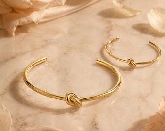 Peut inclure: Deux bracelets joncs en métal doré, chacun présentant un nœud central. Les bracelets ont une finition lisse et polie ainsi qu'une forme circulaire ouverte. Le plus grand bracelet est placé au premier plan, tandis que le plus petit est derrière.