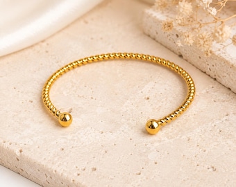 Könnte beinhalten: Ein goldfarbenes Armband in Manschettenform mit einer gedrehten, seilartigen Textur entlang des gesamten Reifs. Das Armband ist offen gestaltet und schließt an beiden Enden mit einer glatten, polierten Kugel ab, was ein schlichtes Design ergibt.