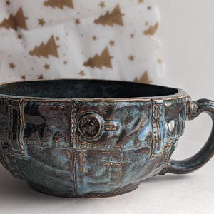 Peut inclure: Une tasse en céramique artisanale avec un design texturé unique. La tasse présente une palette de couleurs bleu foncé et marron avec une anse. L'extérieur présente un motif en relief détaillé. La tasse est adaptée au café ou au thé.