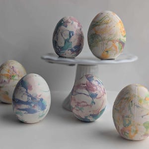 May include: Six decorative Easter eggs with a marbled design in shades of blue, pink, green, and yellow. Two eggs are displayed on a white cake stand, while the others are arranged on a white surface.