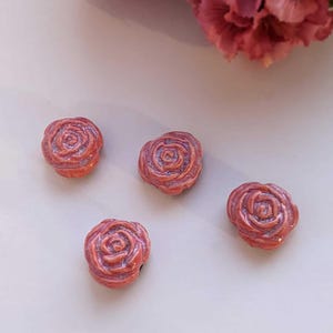 May include: Four coral-coloured rose-shaped beads with detailed petal designs. The beads are arranged on a light background, with a blurred pink flower in the upper right corner. These beads are ideal for jewellery making.