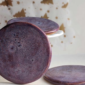 Peut inclure: Ensemble d'assiettes en céramique faites à la main, de couleur violet foncé avec une texture mouchetée. Les assiettes sont rondes, avec un bord légèrement surélevé, et varient en position, certaines empilées sur un support blanc. Le fond présente un motif festif.