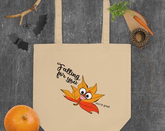 May include: A beige tote bag with a cartoon orange and red leaf with a face and the text "Falling for you" and "you're great".