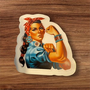 May include: A sticker featuring a woman with dark hair, wearing a red and white polka dot bandana, blue shirt, and turquoise jewelry. She is flexing her bicep with a determined expression. The sticker is on a wooden background.