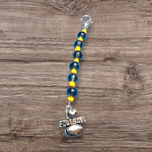 Op de afbeelding: Een blauwe en gele kralen sleutelhanger met een zilveren bedel waarop "Ik hou van voetbal" staat en een voetbalontwerp heeft.