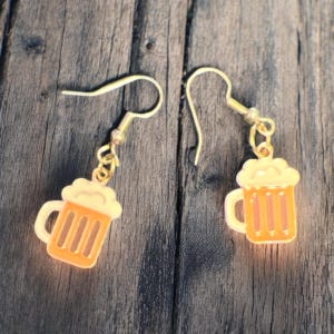 May include: A pair of gold-colored dangle earrings. Each earring features a small, orange beer mug charm with white foam and vertical stripes. The earrings are designed for pierced ears.