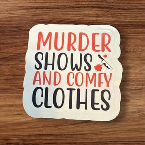 May include: White sticker with black text that reads "Murder Shows and Comfy Clothes". A red knife with blood splatters is next to the word "Shows".