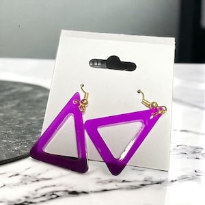 May include: A pair of purple triangle earrings with a gold hook closure. The earrings are translucent and have a gradient effect, fading from a darker purple to a lighter purple.