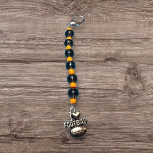Op de afbeelding: Een kralen sleutelhanger met een zilveren sluiting. De sleutelhanger heeft een streng donkerblauwe en oranje kralen. Een zilveren bedel met een voetbal, hart en de woorden "I LOVE FOOTBALL" hangt aan de kralen.