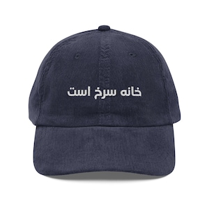 May include: Navy blue corduroy baseball cap with the white text "khaneh sorkh ast" embroidered on the front. The cap has a curved brim and an adjustable strap at the back.